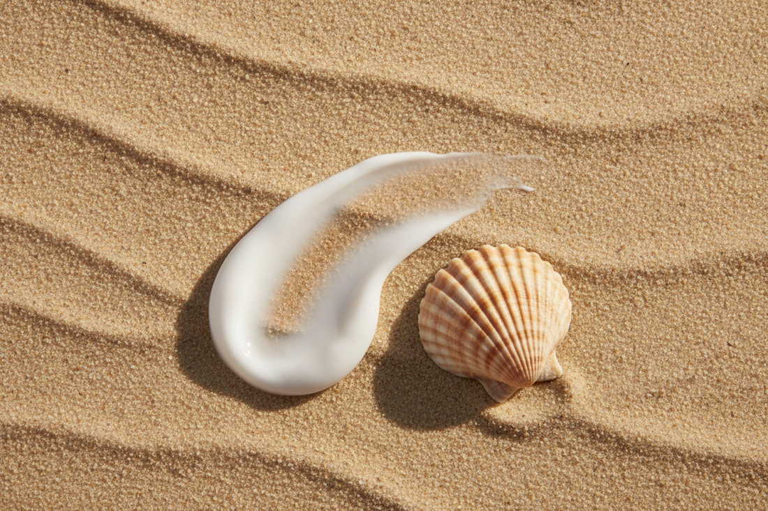 Swirl of white mineral sunscreen cream on warm sand next to a seashell, showing an invisible no white cast finish