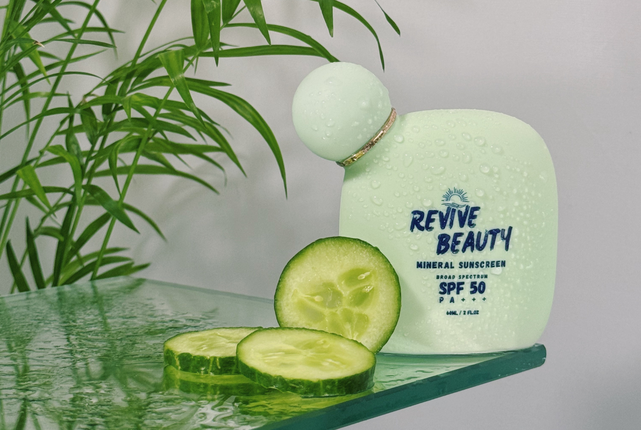 Revive Beauty mineral sunscreen SPF 50 bottle with water droplets on a glass shelf beside fresh cucumber slices and greenery